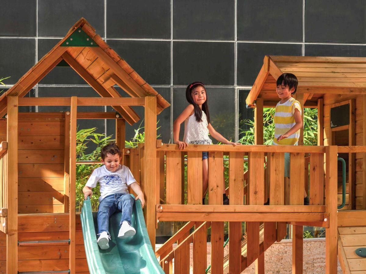Grand Mercure Dubai City Exterior foto The photo shows three children playing on a wooden playground structure. There is a slide on which a young boy is sitting, preparing to slide down. Two other children, a boy and a girl, are standing on a raised platform of the playground. The backgro