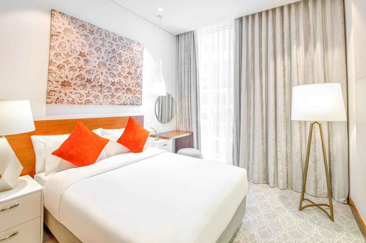 Grand Mercure Dubai City Exterior foto The photo shows a modern hotel room featuring a comfortable queen-sized bed with white linens and orange accent pillows. There is a decorative wall art piece above the bed, adding a stylish touch to the room. A round mirror is mounted on the wall abo