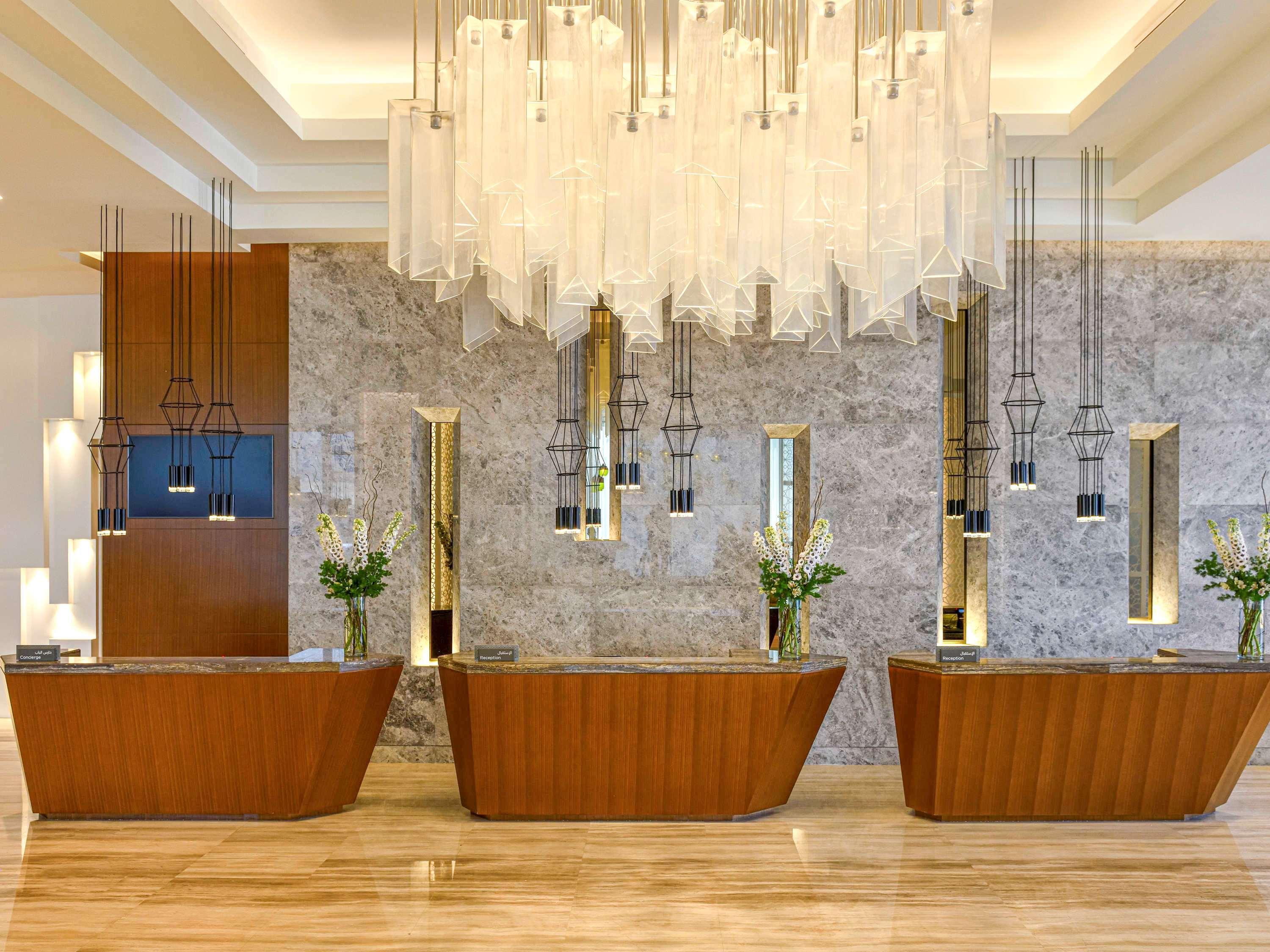 Grand Mercure Dubai City Exterior foto The photo displays a modern hotel reception area characterized by a sleek and stylish design. In the foreground, there are three wooden reception desks that have a smooth, curved shape. Behind the desks, the wall features a mix of marble and decorati
