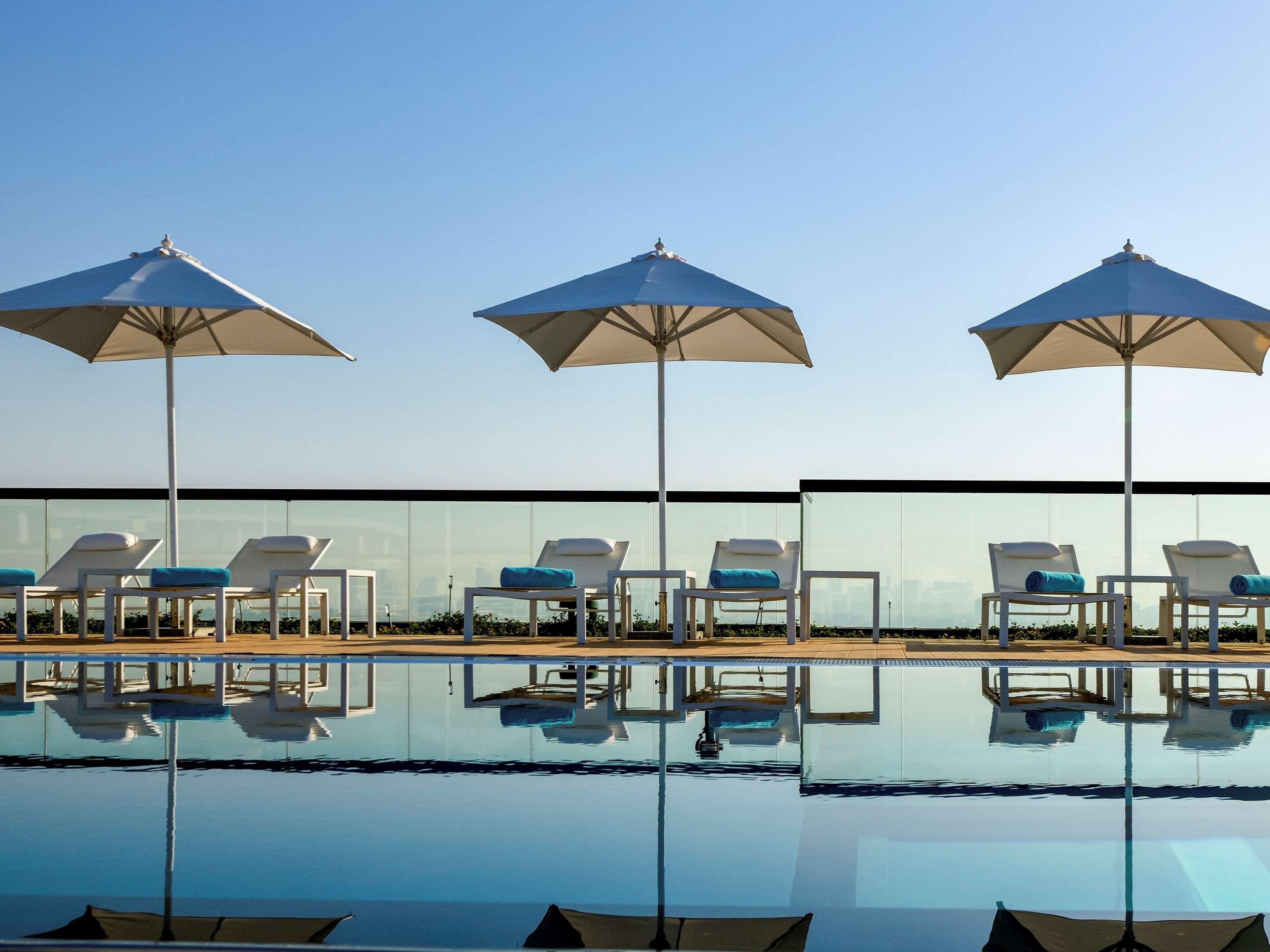 Grand Mercure Dubai City Exterior foto The photo shows a serene poolside scene featuring a series of white lounge chairs with blue cushions positioned alongside a swimming pool. Each chair is shaded by a large, white umbrella. The pool reflects the bright blue sky above, creating a tranqu