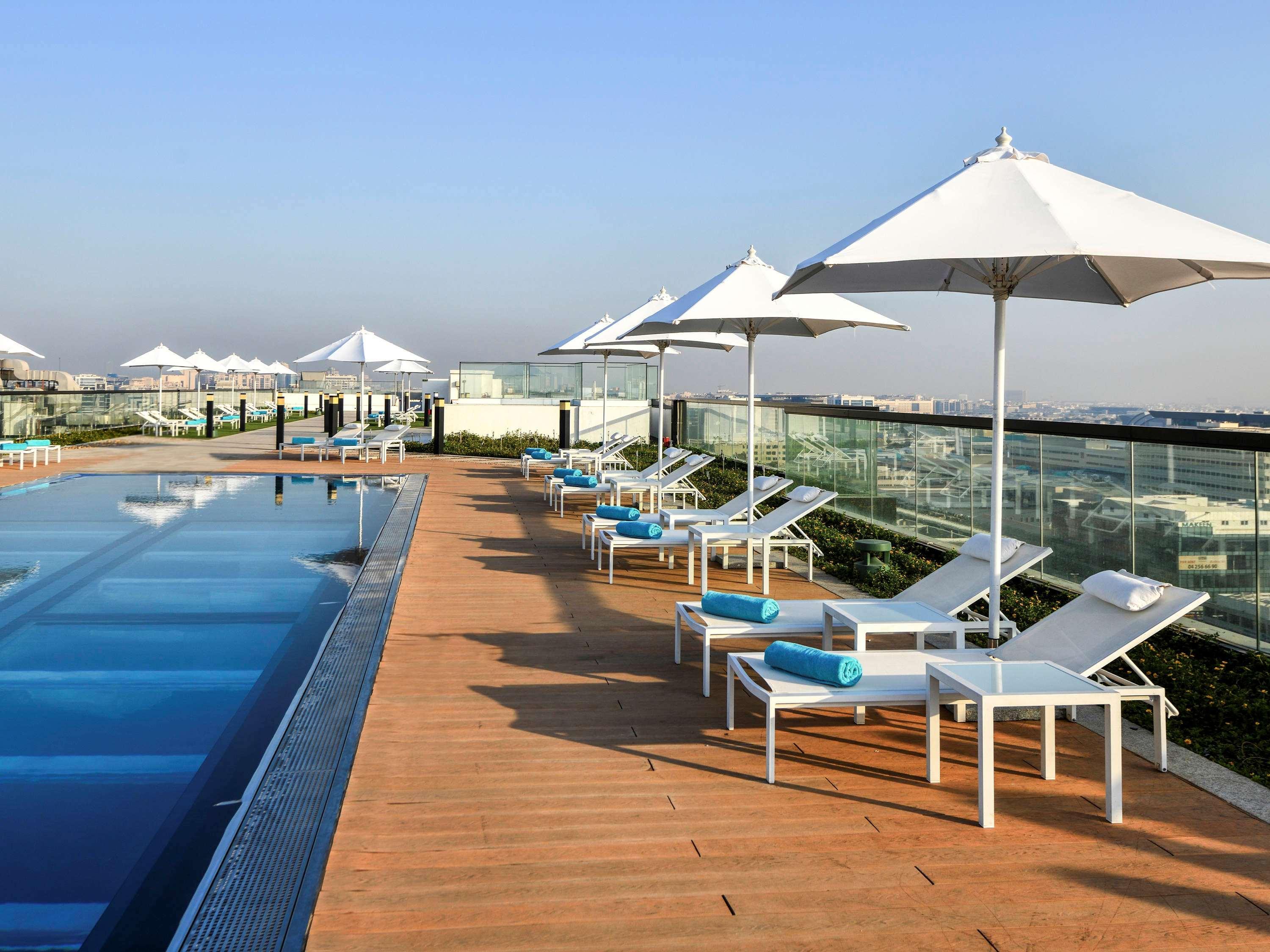 Grand Mercure Dubai City Exterior foto The photo shows a rooftop pool area featuring an infinity pool with clear blue water. Surrounding the pool are several white lounge chairs, each with a light blue cushion. Large white umbrellas provide shade for the loungers. The deck is made of wood