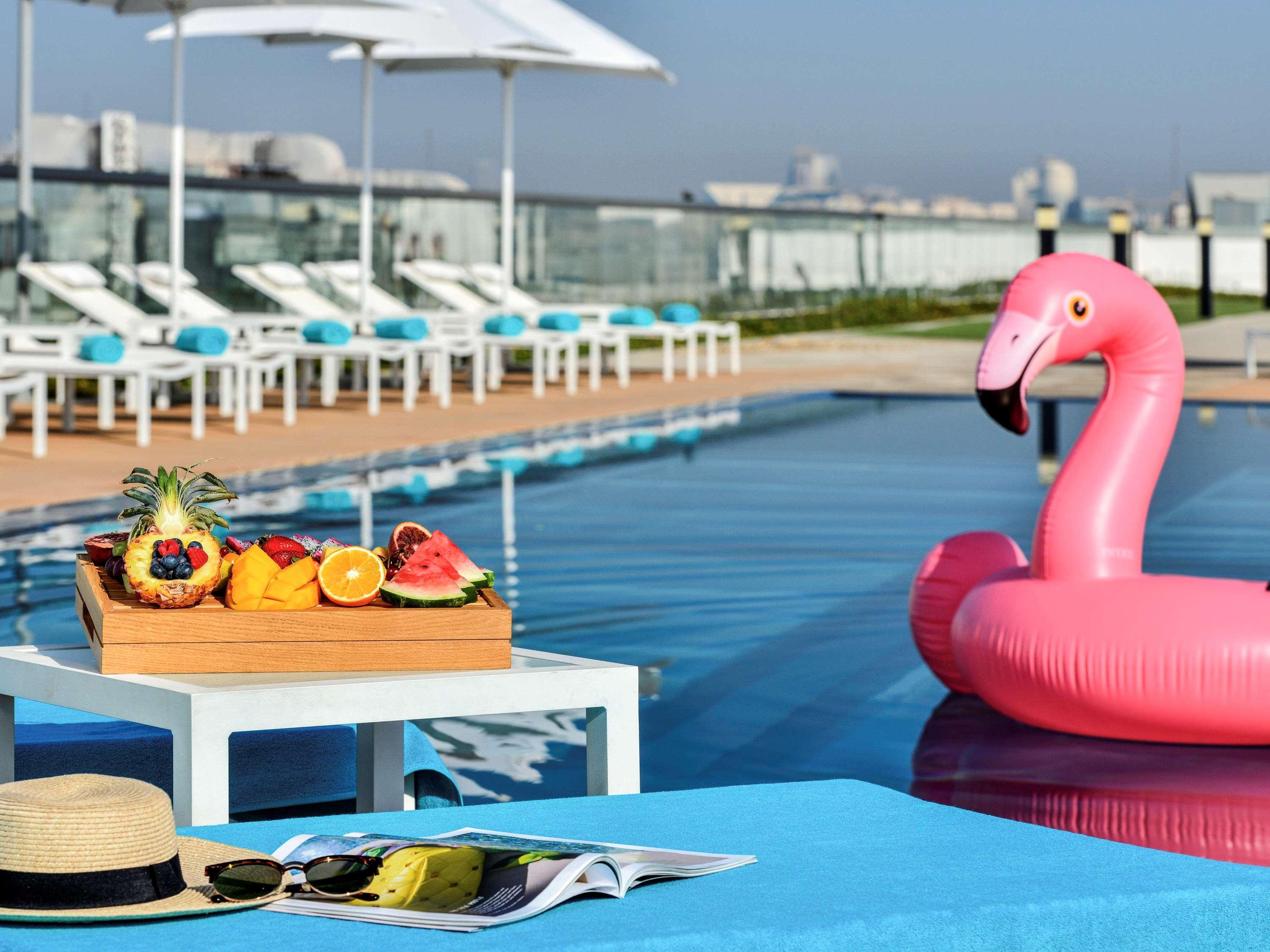 Grand Mercure Dubai City Exterior foto The photo depicts a relaxing poolside scene. In the foreground, there's a small white table with a wooden tray filled with a variety of colorful fruits, including berries, a slice of melon, and citrus fruits. Nearby, there's a beach hat and a magazin