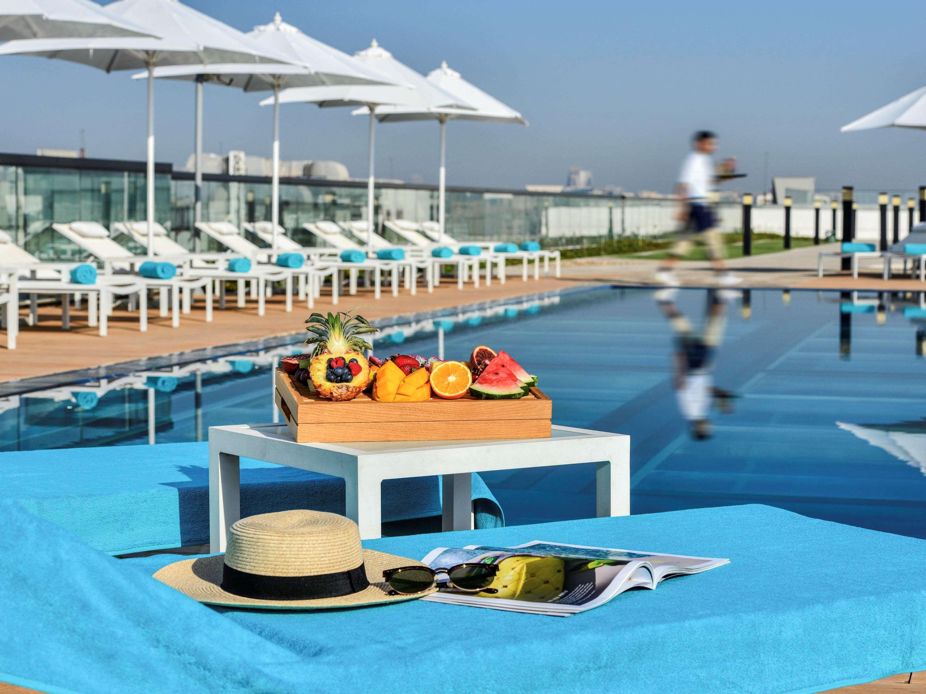 Grand Mercure Dubai City Exterior foto The photo displays a serene poolside scene. In the foreground, there is a table with a wooden tray filled with fresh fruits, including various colorful options. Beside the table, there is a wide-brimmed straw hat and a magazine resting on a light blu