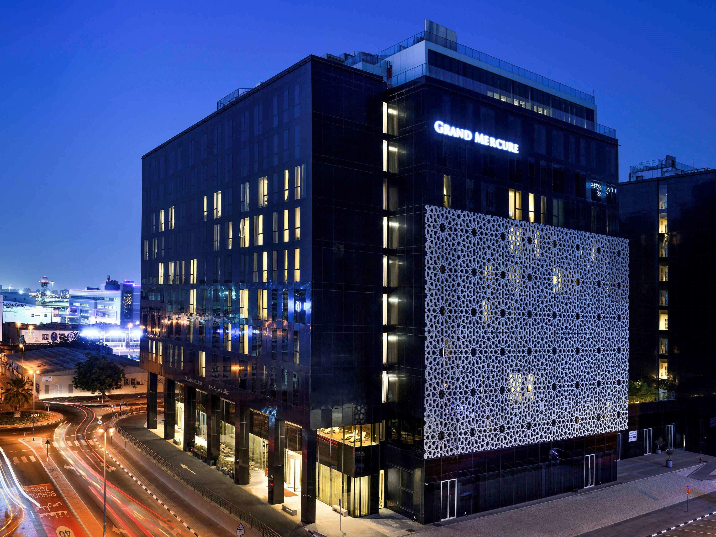 Grand Mercure Dubai City Exterior foto The photo shows a modern building at night, featuring a sleek, contemporary design. The building has a large facade with a distinctive patterned wall on one side. Illuminated signs display the name "Grand Maison." Surrounding the building, there are 