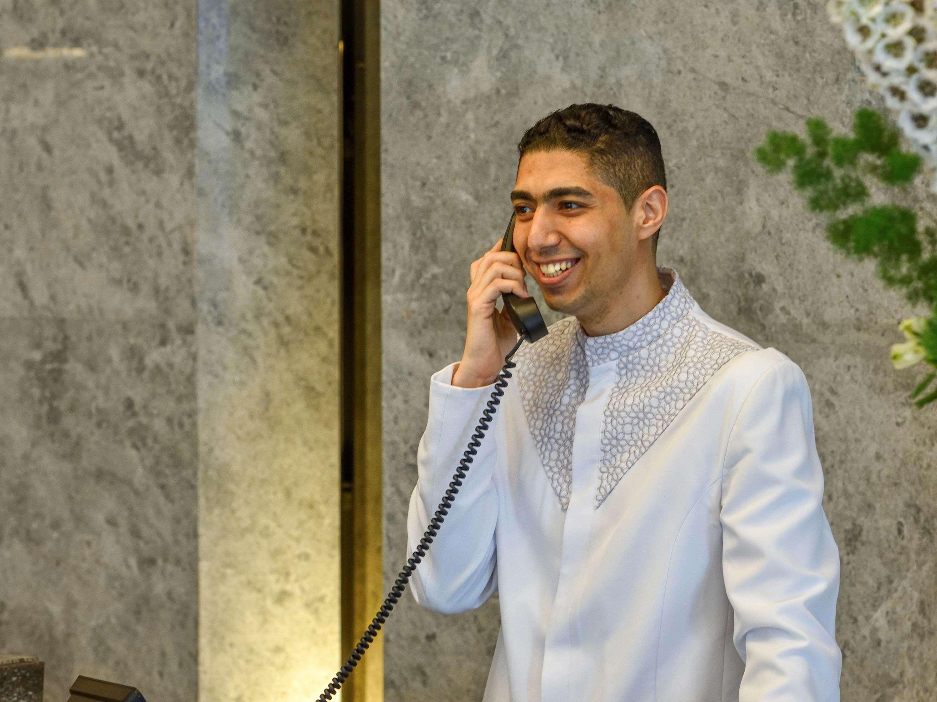 Grand Mercure Dubai City Exterior foto The photo shows a young man dressed in a formal white outfit with decorative details around the collar. He is smiling and is speaking on a vintage-style telephone. The background appears to have a marble texture, and there are some plants or floral a