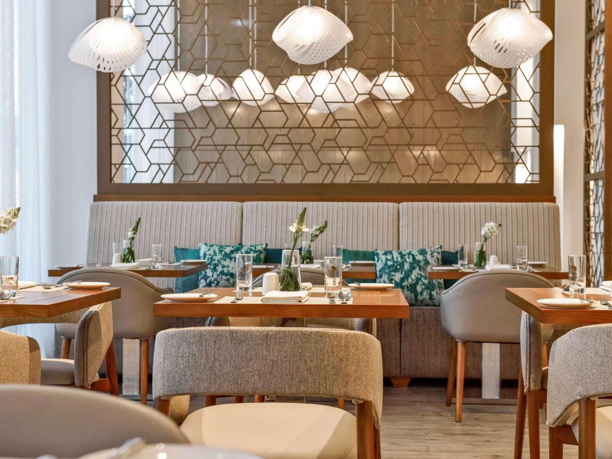 Grand Mercure Dubai City Exterior foto The photo shows a modern restaurant interior. It features dining tables with wooden tops and upholstered chairs. The ambiance is enhanced by pendant lights hanging from the ceiling, which provide soft illumination. The back wall is adorned with a dec
