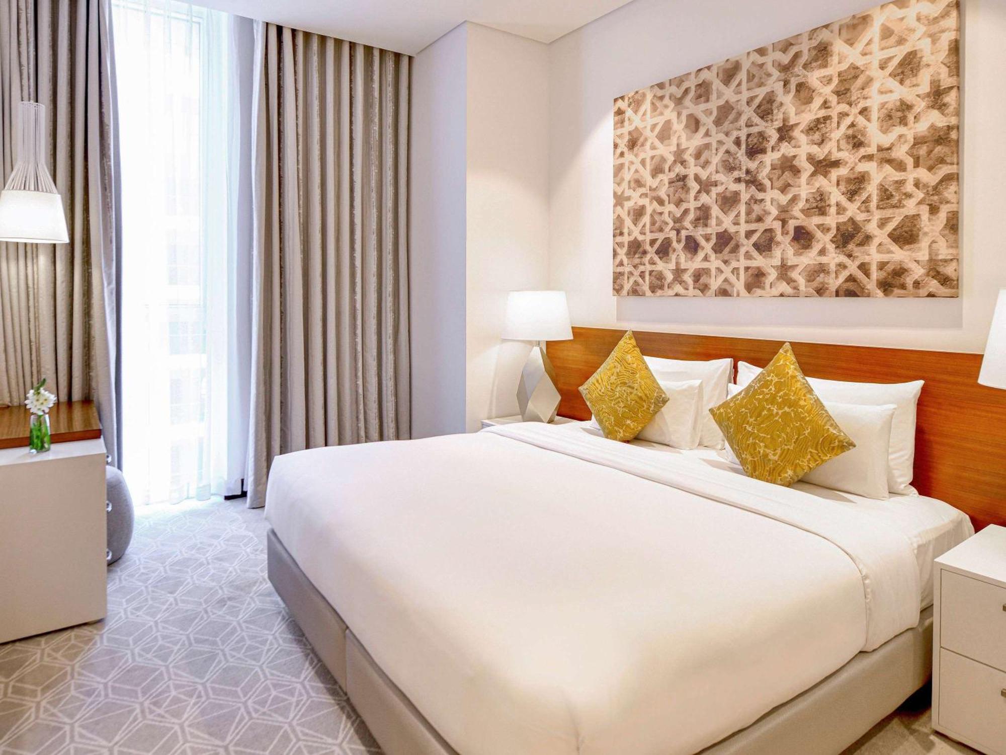 Grand Mercure Dubai City Exterior foto The photo shows a modern hotel room featuring a large bed with white linens and decorative yellow pillows. There are bedside tables on either side of the bed with lamps. The room has large windows with light-colored curtains, allowing natural light t